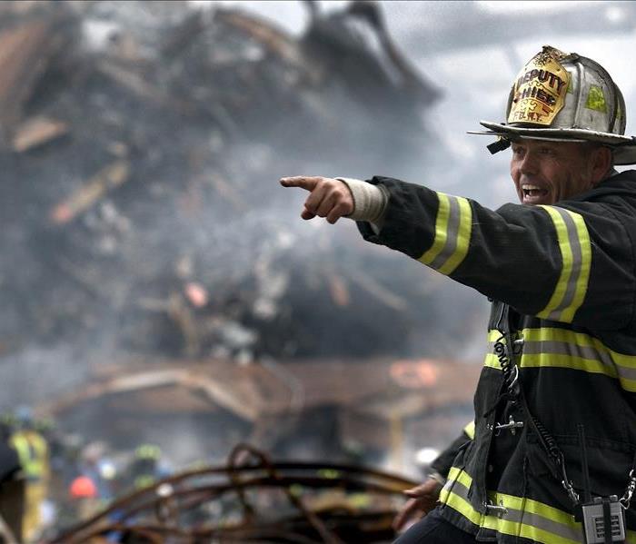 A deputy fire chief in protective gear stands in front of the scene of a disaster, pointing and shouting an instruction.