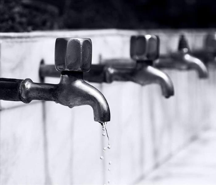 A row of stainless steel water faucets set in a grey brick wall, with one faucet emitting a few clear droplets of water.
